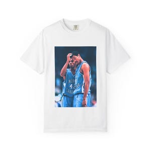 May include: White t-shirt featuring a graphic of two basketball players in blue jerseys. The players are in a moment of reflection. The jerseys have the text "NORTH CAROLINA" and the number "42".