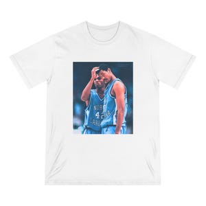May include: White t-shirt featuring a printed image of two basketball players in light blue jerseys. The jerseys display the text "North Carolina" and the number "42". The players are in a close-up shot, with one touching his head.