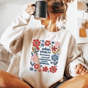 May include: Cream-colored sweatshirt with a floral design and the text "when cruelty becomes normal compassion seems radical." The design features red, blue, and pink flowers. A black mug is held up to the face.