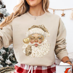 May include: A beige sweatshirt featuring a Santa Claus graphic with gold accents and a leopard print hat. The person is wearing red and white plaid pajama pants and holding a white mug with a Santa illustration.
