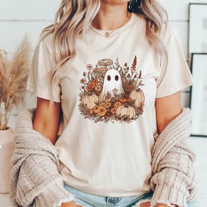 May include: Beige t-shirt featuring a whimsical illustration of a ghost surrounded by pumpkins, flowers, and mushrooms in autumnal colors. The design has a vintage, boho aesthetic. The shirt is a casual, everyday wear item.