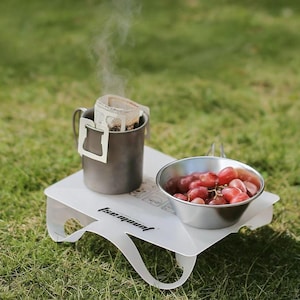 Puede incluir: Una mesa de camping blanca y plegable sostiene una taza de metal con un filtro de café, emitiendo vapor, y un pequeño cuenco de metal lleno de uvas rojas. La mesa está sobre hierba.