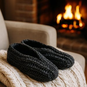 May include: A pair of dark gray knitted slippers rests on a cream-colored cable-knit blanket. The slippers are made of a thick, textured yarn. A cozy fireplace with a warm glow is in the background, suggesting comfort and relaxation.