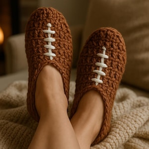 May include: Brown crochet slippers designed to resemble footballs, with white stitching detail. The slippers are shown on a person's feet, resting on a beige knitted blanket. The cozy footwear is perfect for relaxing at home.