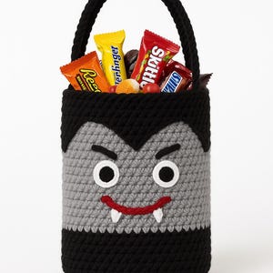 May include: A handmade crochet Halloween candy basket shaped like a vampire. The black and gray basket has a handle and is filled with assorted candy bars. The vampire face has white eyes, a red smile, and two white fangs.