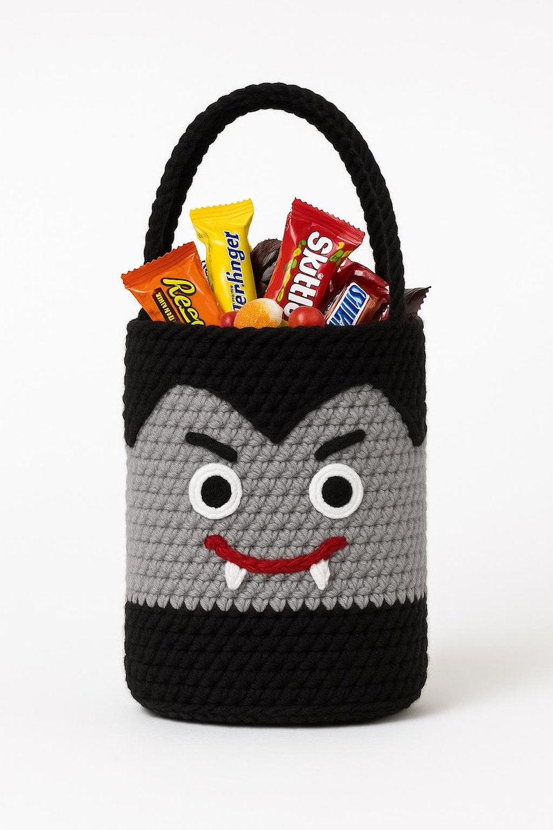 May include: A handmade crochet Halloween candy basket shaped like a vampire. The black and gray basket has a handle and is filled with assorted candy bars. The vampire face has white eyes, a red smile, and two white fangs.