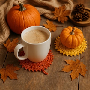 May include: A white mug of coffee rests on an orange crocheted leaf coaster. The image includes a small orange pumpkin, autumn leaves, and a yellow crocheted coaster with a small pumpkin. The scene evokes a cosy autumn atmosphere.