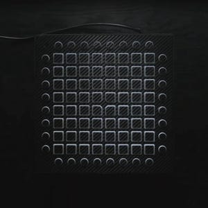 May include: Black electronic music production device with a carbon fibre texture. It features a grid of square and circular buttons, and a cable extending from the top. The buttons are white and the device is set against a dark background.