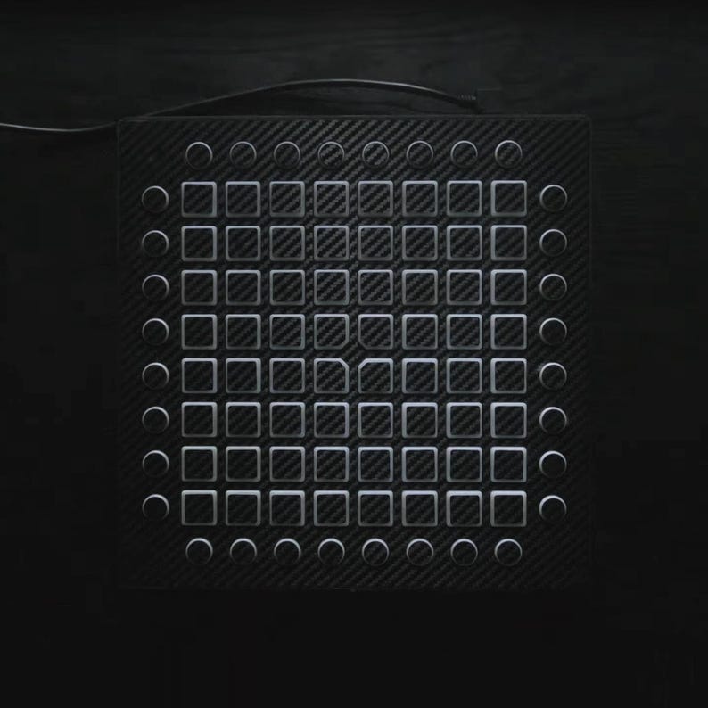 May include: Black electronic music production device with a carbon fibre texture. It features a grid of square and circular buttons, and a cable extending from the top. The buttons are white and the device is set against a dark background.