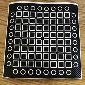 May include: A black and white sticker featuring a grid of white squares and circles. The sticker has a carbon fibre texture and is displayed on a wooden surface.