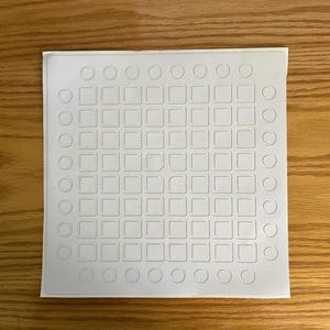 May include: A white, square sheet featuring a grid of small squares and circles. The sheet has a textured surface and is set against a wooden background. The design is evenly spaced, creating a symmetrical pattern.