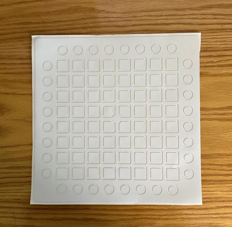 May include: A white, square sheet featuring a grid of small squares and circles. The sheet has a textured surface and is set against a wooden background. The design is evenly spaced, creating a symmetrical pattern.
