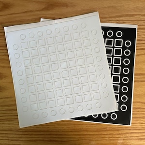 May include: Two sticker sheets, one white with embossed squares and circles, the other black with white shapes. The stickers feature a grid pattern and are on a wooden surface.