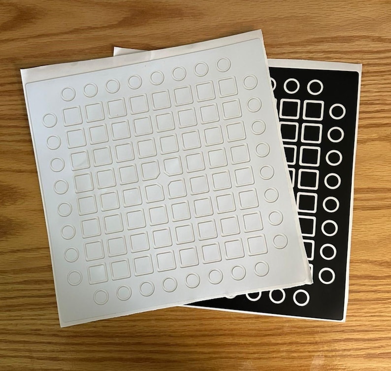 May include: Two sticker sheets, one white with embossed squares and circles, the other black with white shapes. The stickers feature a grid pattern and are on a wooden surface.