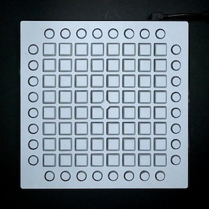 May include: A square, light grey metal plate featuring a grid of square and circular indentations. The plate has a series of square cutouts in the centre, surrounded by circular cutouts along the edges. A black cable extends from the top.