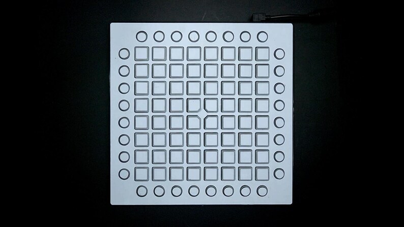 May include: A square, light grey metal plate featuring a grid of square and circular indentations. The plate has a series of square cutouts in the centre, surrounded by circular cutouts along the edges. A black cable extends from the top.