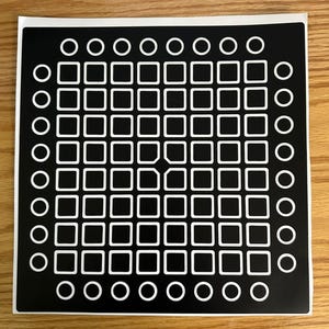 May include: A black sticker with a grid pattern of white squares and circles. The design features a central array of 64 squares, surrounded by a border of circles. The sticker is on a wooden surface.