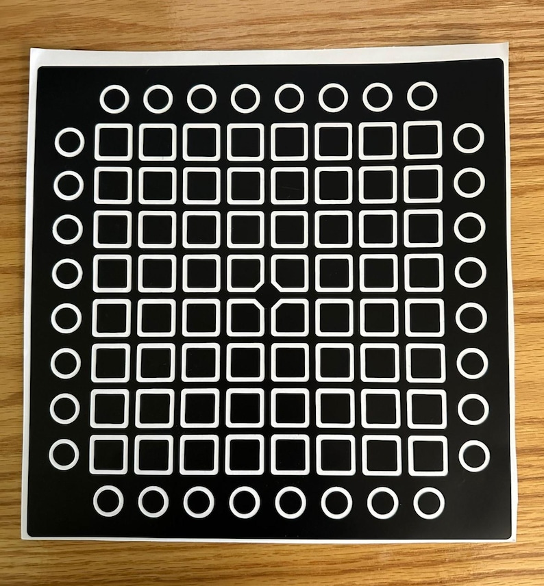May include: A black sticker with a grid pattern of white squares and circles. The design features a central array of 64 squares, surrounded by a border of circles. The sticker is on a wooden surface.
