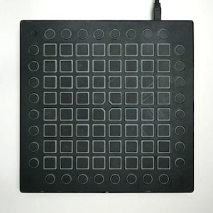 May include: A black electronic music production device with a grid of square and circular buttons. The device has a rectangular shape and a cable extending from the top. The buttons are arranged in a grid pattern.