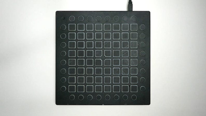 May include: A black electronic music production device with a grid of square and circular buttons. The device has a rectangular shape and a cable extending from the top. The buttons are arranged in a grid pattern.