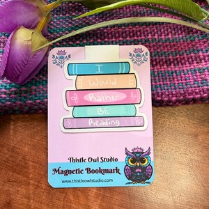 May include: A pink magnetic bookmark with stacked book illustrations in blue, peach, pink, and teal. The text "I Would Rather BE Reading" is on the books. The bookmark has a Thistle Owl Studio logo with a purple owl.
