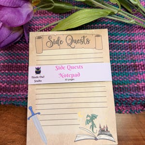 May include: A notepad with the text "Side Quests" on the top and a pink band that says "Side Quests Notepad 30 pages". The notepad has a sword, a dragon, a castle, and an open book illustration. The notepad is on a wooden surface.