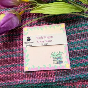 May include: Pink sticky notes with a floral border and the text "Book Dragon Sticky Notes" and "30 pages." A green dragon illustration sits on a stack of books. The notes are on a colorful woven surface with purple flowers.