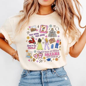 Op de afbeelding: Een crèmekleurig t-shirt met een kleurrijke grafische print met diverse illustraties, waaronder een rode scooter, een koffer, een paspoort, een afstudeerhoed en twee figuren. De tekst luidt "HEY NOW! HEY NOW!", "Sing to me PAOLO" en "THIS IS WHAT DREAMS ARE MADE OF".