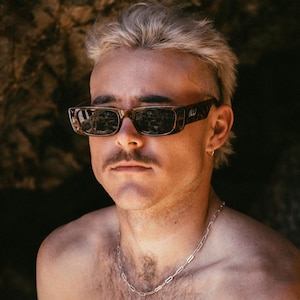 Op de afbeelding: Een jonge persoon met lichtblond haar, die een schildpadzonnebril en een zilveren ketting draagt. De persoon heeft een tatoeage van een paraplu op zijn schouder.