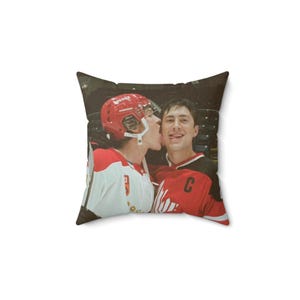 May include: Square pillow featuring a photograph of two hockey players in red and white uniforms. One player is kissing the other on the cheek. The pillow has a dark background.