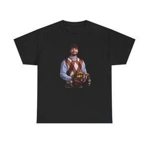 May include: Black t-shirt featuring a person wearing a blue shirt and a brown and white argyle sweater, holding a brown and gold object. The person is smiling. The t-shirt is a basic crew neck style.