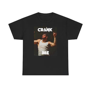 May include: Black t-shirt with a graphic of a person wearing a white tank top and red face paint. The words "CRANK ME" are printed in white above and below the image.