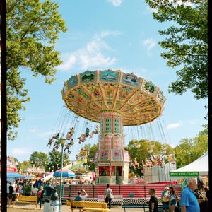 May include: A colorful carousel with people riding in the swings. The carousel is in a park setting with trees and a sign that says "Free Kids Crafts".