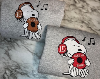May include: Two gray sweatshirts featuring an embroidered Snoopy character wearing headphones with the number "1D" on them. Snoopy is holding a brown record with the word "FOUR" on one and a red record with "TAKE ME HOME" on the other. Musical notes are embroidered above.