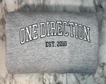 May include: A folded gray sweatshirt with the embroidered text "ONE DIRECTION" in white, outlined in black, arched across the top. Below the text is "EST. 2010" in a smaller, black font.