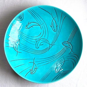 May include: A round, bright turquoise ceramic bowl featuring a series of black line drawings of fish swimming in various directions. The design includes flowing, curved lines that suggest movement and water currents across the bowl's surface.