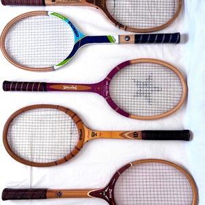 May include: Five vintage wooden tennis rackets with various handle and frame designs. The rackets feature different brand names, including Wilson, Spalding, and Davis. The rackets are arranged on a white surface, showcasing their aged wood and string netting.