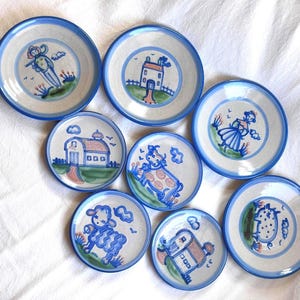 May include: A collection of seven round ceramic plates with a light gray base and blue rims. Each plate features hand-painted blue, green, and brown illustrations including a person, a house, a barn, a cow, a sheep, and other rural scenes.