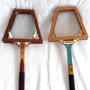 May include: Two vintage wooden tennis rackets with wire mesh netting. One racket has a dark brown grip and the other has a black grip. The rackets have a light brown wooden frame and a decorative design on the handle.