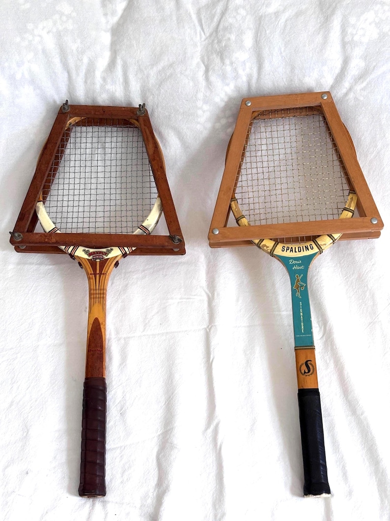 May include: Two vintage wooden tennis rackets with wire mesh netting. One racket has a dark brown grip and the other has a black grip. The rackets have a light brown wooden frame and a decorative design on the handle.