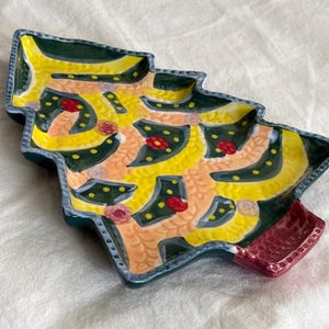 May include: A ceramic Christmas tree-shaped dish with a yellow interior, decorated with green, red, and yellow accents. The dish has a dark green border and a red base. The design is hand-painted and features a festive, whimsical style.