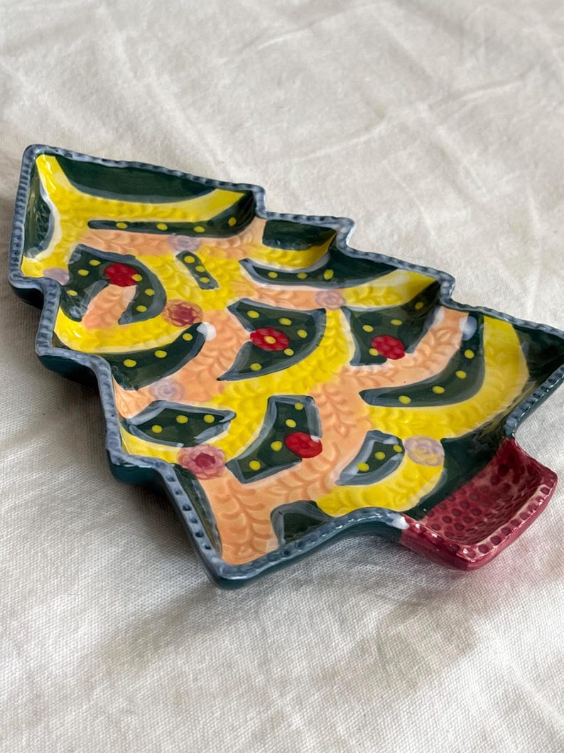 May include: A ceramic Christmas tree-shaped dish with a yellow interior, decorated with green, red, and yellow accents. The dish has a dark green border and a red base. The design is hand-painted and features a festive, whimsical style.
