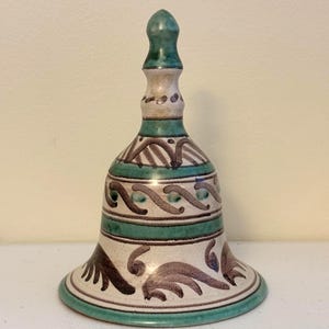 May include: Ceramic bell with a green, white, and brown color scheme. The bell features a decorative design with a green handle and a flared base. The bell's surface is adorned with intricate patterns and a glossy finish.