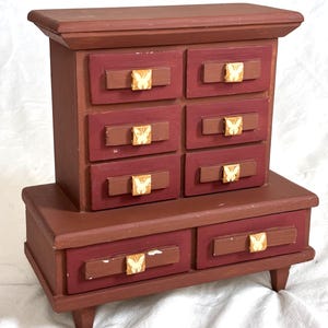 May include: A small, wooden jewelry box with a dark red finish. It features six small drawers on top and two larger drawers below. Each drawer has a brown handle with a butterfly design. The box sits on short, angled legs.