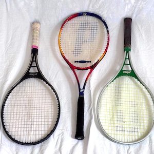 May include: Three vintage Prince tennis rackets. One black racket with a white grip, a red, yellow, and blue racket with a black grip, and a green racket with a brown grip. All rackets have white stringing and the Prince logo.