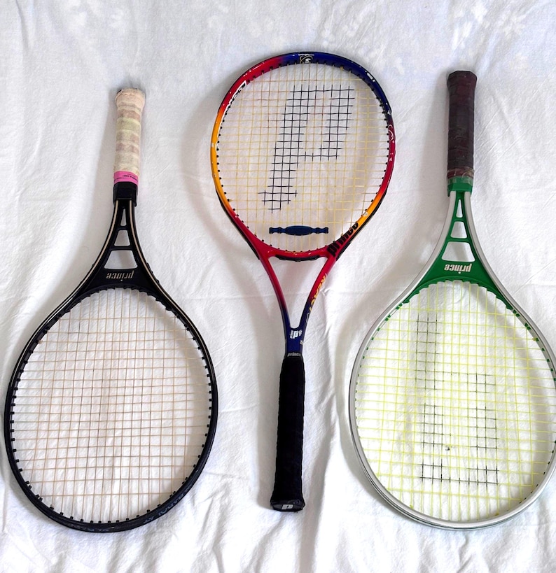 May include: Three vintage Prince tennis rackets. One black racket with a white grip, a red, yellow, and blue racket with a black grip, and a green racket with a brown grip. All rackets have white stringing and the Prince logo.