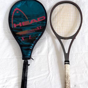 May include: Two tennis rackets, one with a teal and black cover featuring the word "HEAD" in pink. The other racket is gray with a white grip. The rackets are on a white surface.