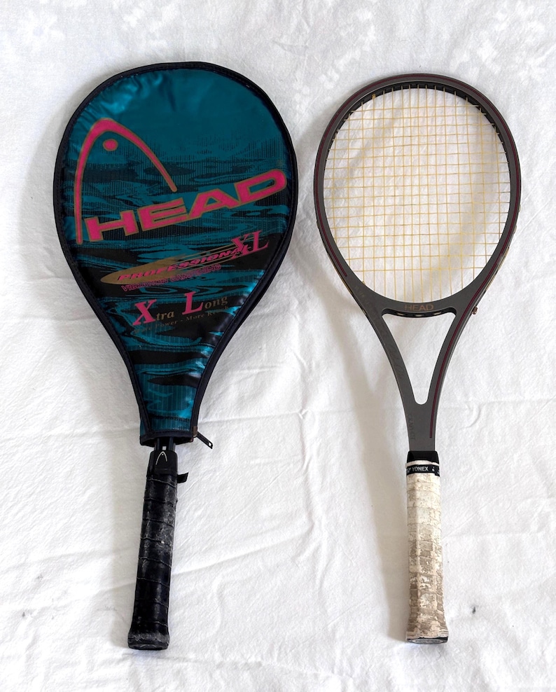 May include: Two tennis rackets, one with a teal and black cover featuring the word "HEAD" in pink. The other racket is gray with a white grip. The rackets are on a white surface.