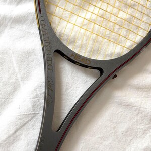 May include: A gray HEAD Competition Edge tennis racket with yellow strings. The racket has red accents and the text "Graphite" and "Carl-Uwe" is visible. The racket is on a white surface.