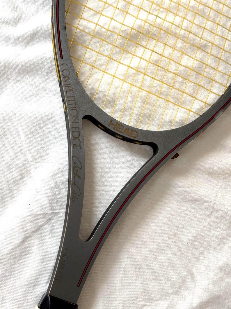 May include: A gray HEAD Competition Edge tennis racket with yellow strings. The racket has red accents and the text "Graphite" and "Carl-Uwe" is visible. The racket is on a white surface.